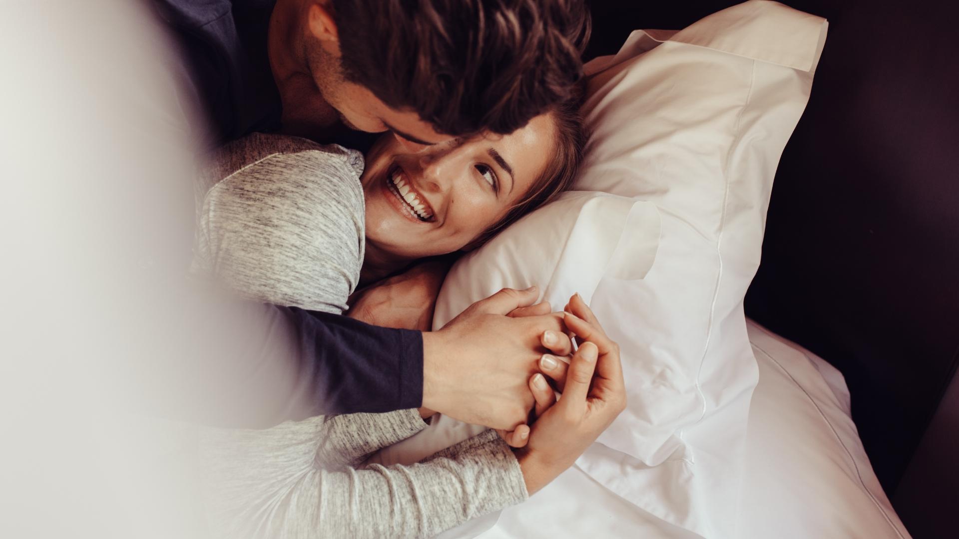 A happy couple cuddling in bed, smiling and holding hands.