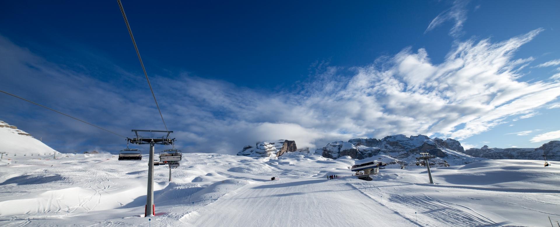 Verschneite Landschaft mit Sessellift und Bergen im Hintergrund unter blauem Himmel.