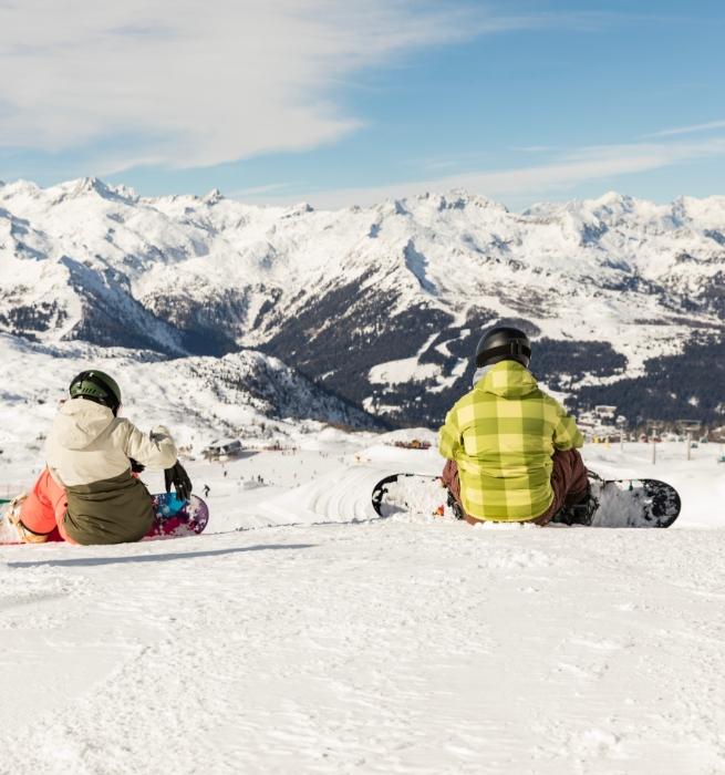 Two snowboarders rest on the snow with a view of snowy mountains.