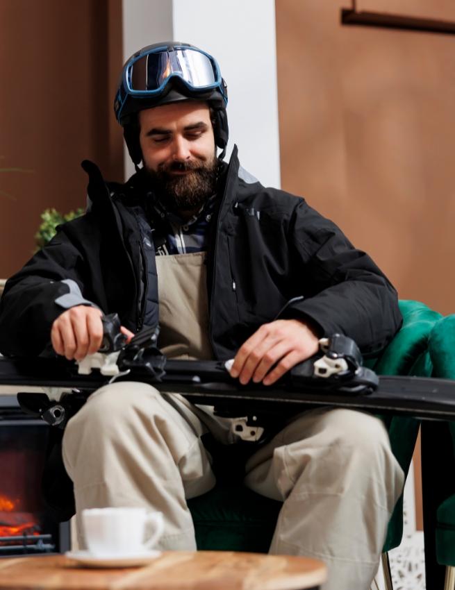 Skier with helmet and goggles prepares gear near the fireplace.